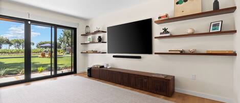 Un soggiorno moderno con una grande TV a schermo piatto fissata al muro, mensole in legno con oggetti decorativi, un mobile console in legno integrato e un'ampia porta scorrevole in vetro che dà su un giardino.