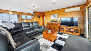 A modern living room with a black leather sofa, wooden cabinets, a TV, and a checkered rug. - Riverside Haven - Turangi Holiday Home (Turangi)