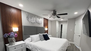 Ein modernes Schlafzimmer mit einem großen Bett, einem Nachttisch, einer Blumenvase, einem Deckenventilator und einem an der Wand montierten Fernseher.