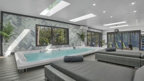 A modern indoor pool area with a hot tub, seating, and large windows offering views of the outdoors.