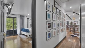 A room with a bed, a wall of framed pictures, a dining area with chairs, and a view of trees through a window.