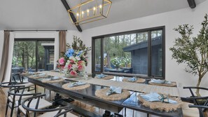 A long dining table with chairs, a vase of flowers, and a view of the outdoors through large windows.