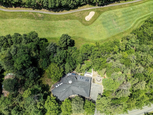 An aerial view of a property with a large building surrounded by dense forest, a winding road, and a well-maintained lawn with a sand bunker.