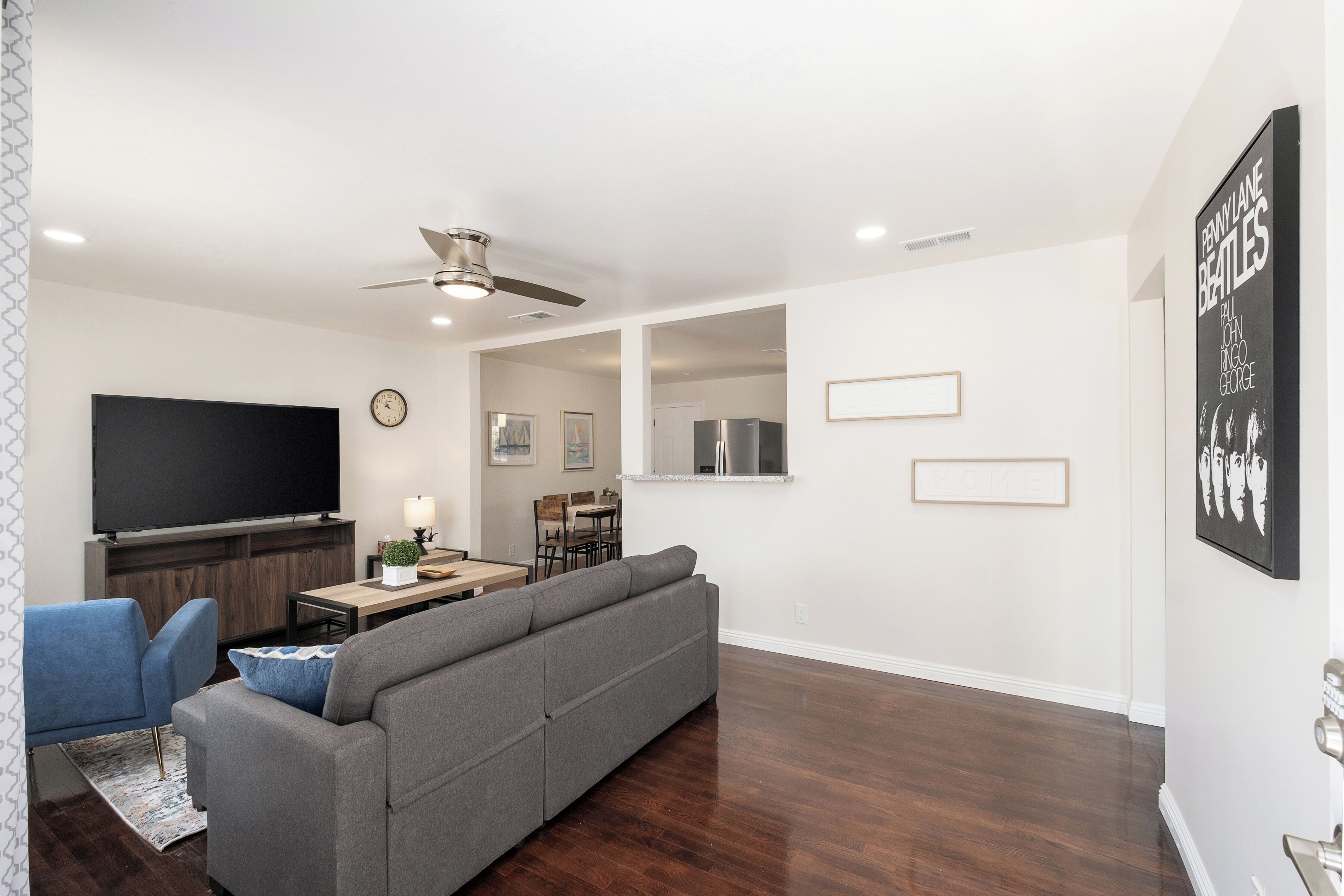 A modern living room with a grey sectional sofa, a flat-screen TV, a ceiling fan, and a dining area with a wooden table and chairs.