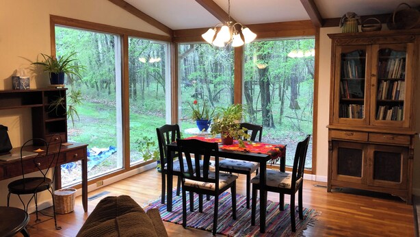A spacious room with large windows offering a forest view, a dining table with chairs, a cozy sofa, and a wooden cabinet with books.