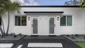 Un edificio moderno con dos puertas identificadas con los números 2 y 3, techo negro y un área de estacionamiento delimitada con bolardos blancos.
