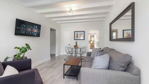 Una sala moderna con un sofá gris, una mesa de centro de madera y una pintura de un paisaje urbano. Hay una zona de comedor con una mesa y sillas de vidrio, y un espejo grande en la pared.