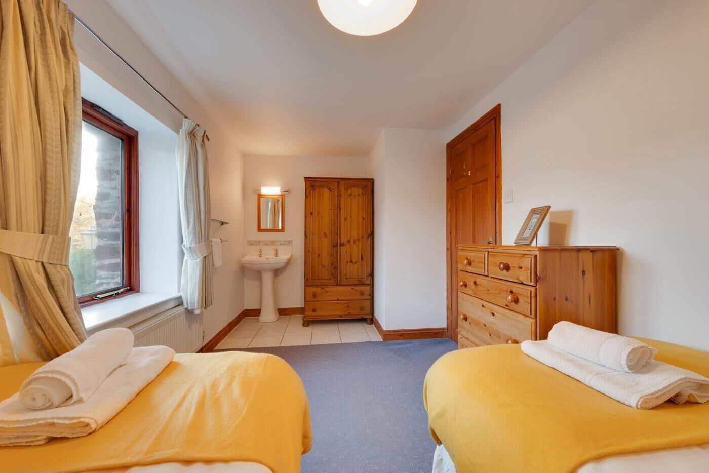 A bedroom with two beds, a wooden wardrobe, a bedside table, a mirror, and a window with curtains.