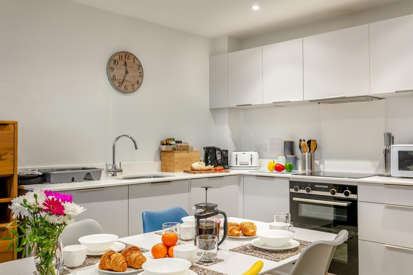 A modern kitchen with a sink, stove, and microwave, a dining table with breakfast items, and a clock on the wall.