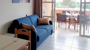 A living room with a blue sofa, a wooden dining table, and chairs. There is a balcony with a table and chairs visible through the glass doors. - T2 apartment with sea view and private parking in Cap Esterel vacation village (Saint-Raphaël)