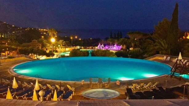 A large, illuminated swimming pool surrounded by lounge chairs and umbrellas, with a view of a hill and buildings in the background. - T2 apartment with sea view and private parking in Cap Esterel vacation village (Saint-Raphaël)