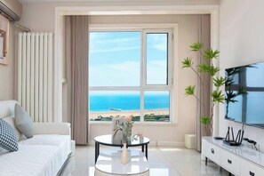 Habitación de hotel moderna con un ventanal grande que ofrece vista a la playa, una cama, un televisor y una mesita con un florero.