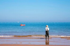 Een strandzicht met iemand die aan de kust staat, een rode opblaasbare luchtbed in het water en een helderblauwe lucht.
