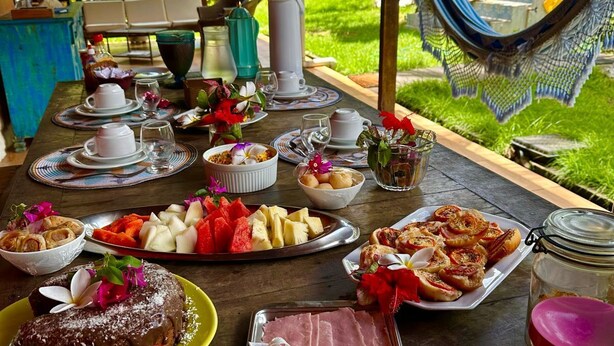 Ein Essbereich im Freien mit einer Auswahl an Speisen und Getränken, darunter Gebäck, Obst und Käse, vor einer üppigen Gartenkulisse.