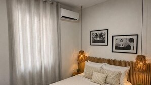 Ein Schlafzimmer mit Bett, Vorhängen, Klimaanlage, gerahmten Bildern und einem Nachttisch mit Lampe.