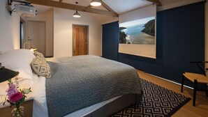 A bedroom with a bed, a nightstand, a lamp, a window with a view of a beach, and a projector mounted on the ceiling.