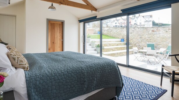 A bedroom with a large bed, a wooden ceiling, and a view of a stone wall and outdoor seating area.