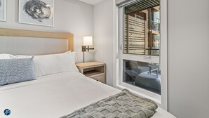 Ein modernes Hotelzimmer mit einem großen Bett, einem Nachttisch mit Lampe, einem Fenster mit Blick ins Innere des Gebäudes und Kunstwerken an der Wand.