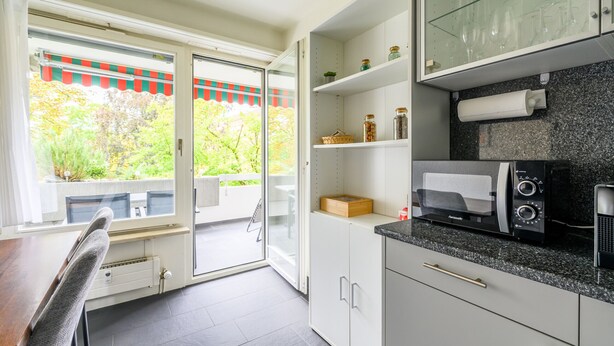 A modern kitchen with a built-in microwave, a glass-fronted cabinet with jars, and a sliding glass door leading to an outdoor seating area. - Marllon Suites – St. Alban Apartments – Apartment Arnold (Basel)