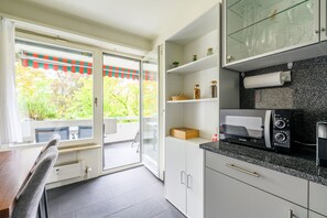 A modern kitchen with a built-in microwave, a glass-fronted cabinet with jars, and a sliding glass door leading to an outdoor seating area. - Marllon Suites – St. Alban Apartments – Apartment Arnold (Basel)