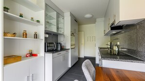 A modern kitchen with white cabinetry, a granite countertop, and a wooden dining table. - Marllon Suites – St. Alban Apartments – Apartment Arnold (Basel)