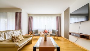 A modern living room with a leather sofa, two armchairs, a glass coffee table, and a flat-screen TV mounted on the wall. - Marllon Suites – St. Alban Apartments – Apartment Arnold (Basel)