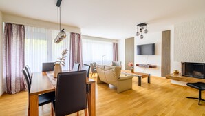 A modern living room with a wooden dining table, chairs, a sofa, and a TV mounted on the wall. - Marllon Suites – St. Alban Apartments – Apartment Arnold (Basel)