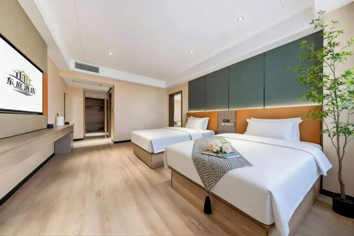 A hotel room with two beds, a large mirror, a plant, and a screen displaying the hotel name.