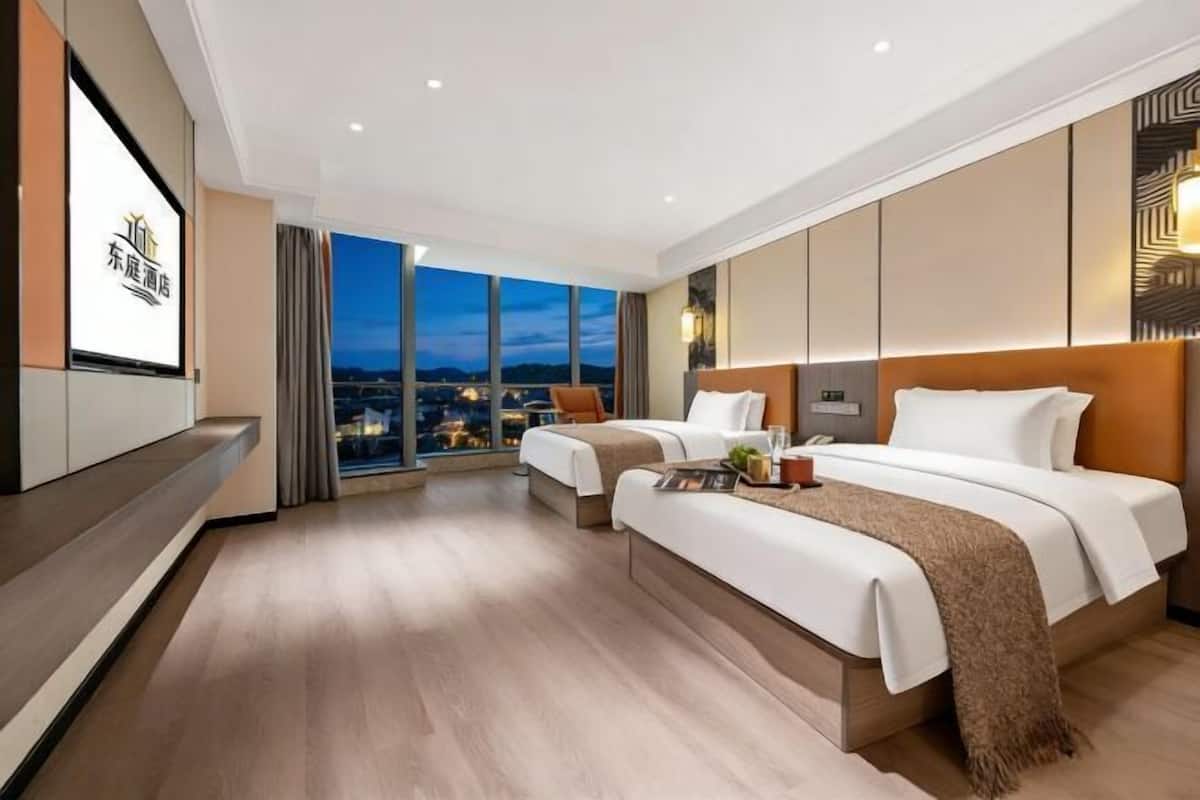 A hotel room with two beds, a large window with a city view, a TV, and a wooden floor.