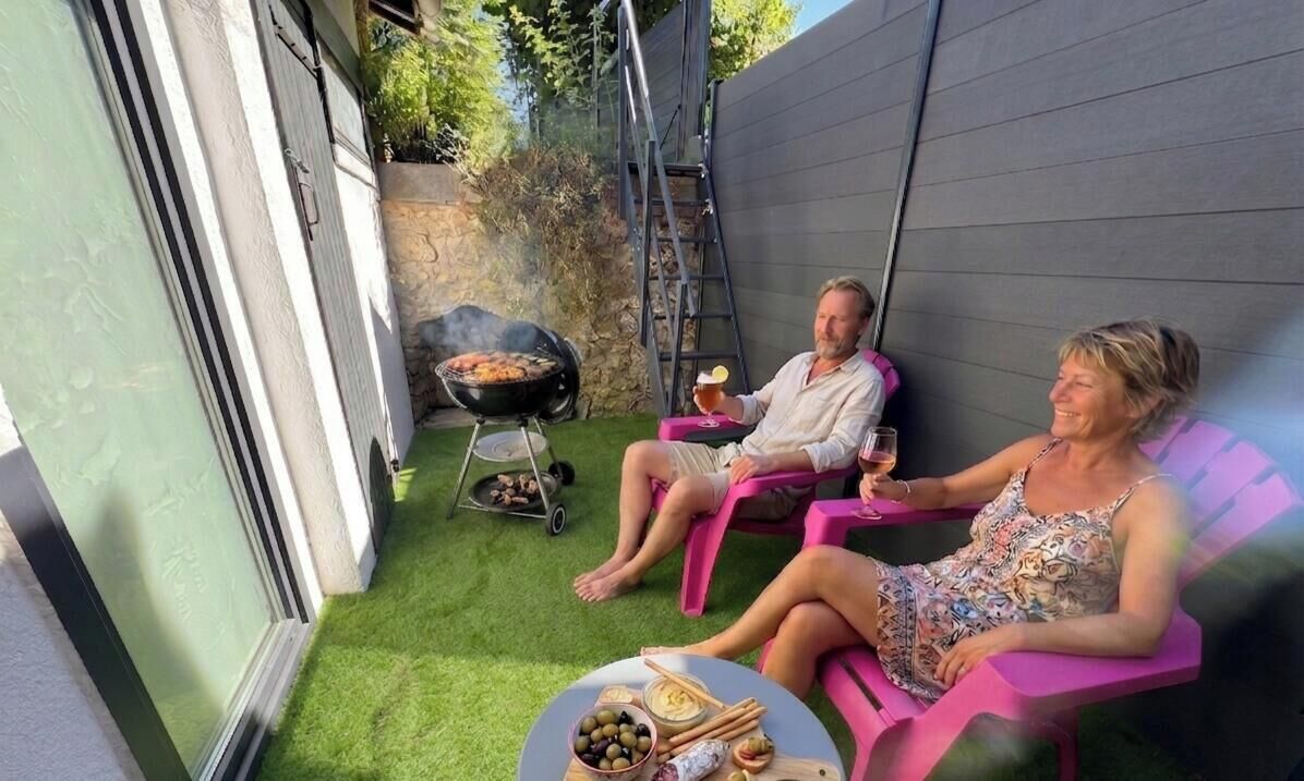 Zwei Personen sitzen draußen auf rosa Stühlen, im Vordergrund steht ein Grill und ein Tisch mit Speisen.
