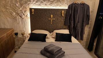Een slaapkamer met een stenen muur, een bed met hoofdeinde, twee zwarte kussens, een opgevouwen handdoek en een houten nachtkastje met een kom en een glas.