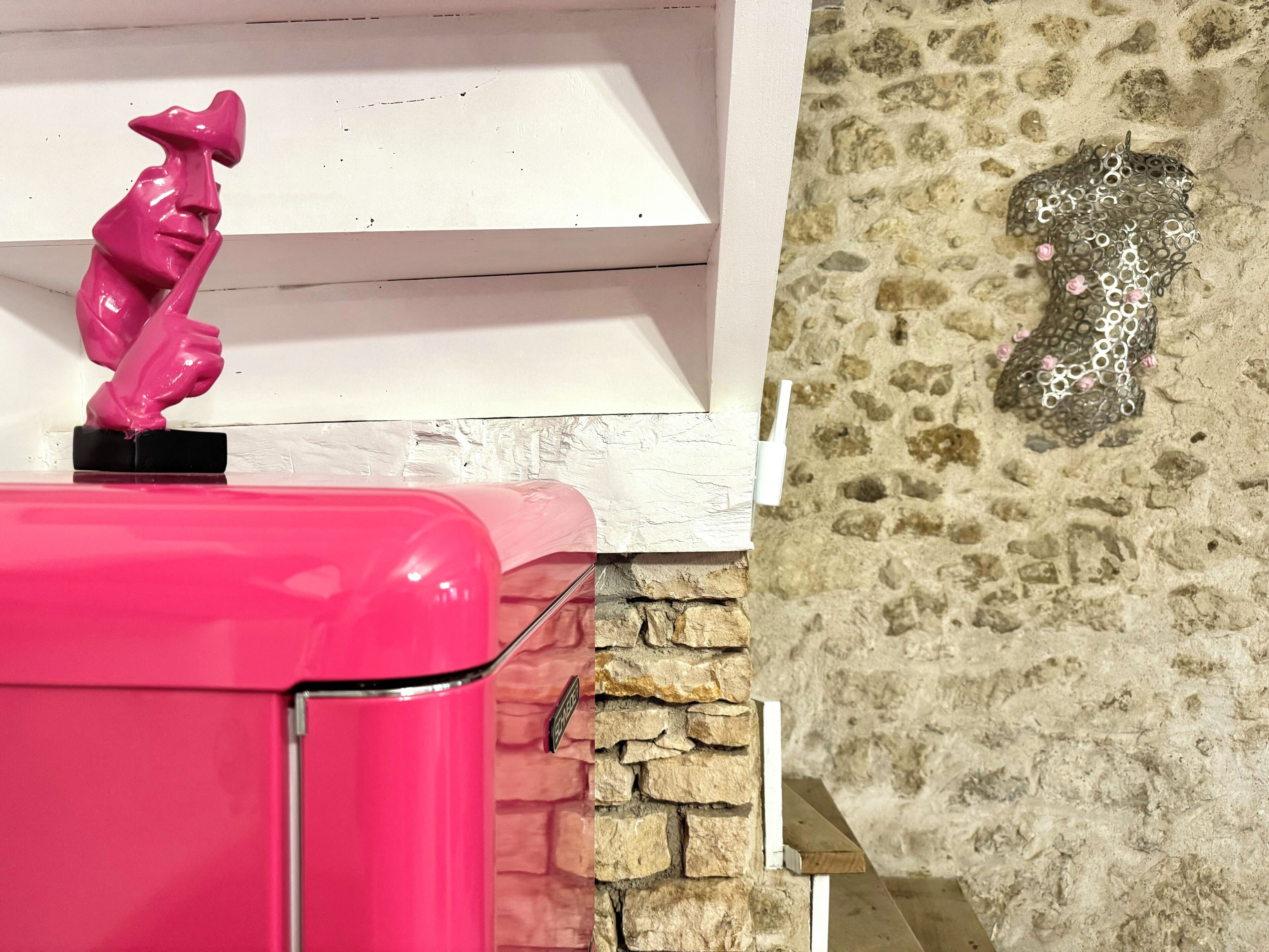 Une valise rose sur une étagère blanche, une sculpture rose décorative et un mur de pierre à motifs texturés.