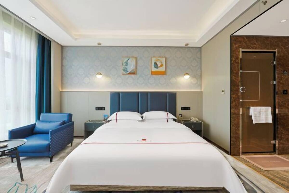 Une chambre d’hôtel avec un lit, un fauteuil bleu, une table de chevet, une commode et une porte sur laquelle est accrochée une serviette.
