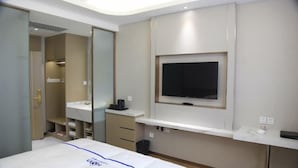 A modern hotel room with a bed, a TV mounted on the wall, a vanity with a mirror, and a small desk area. - Tianxing Hotel (Lingshi)