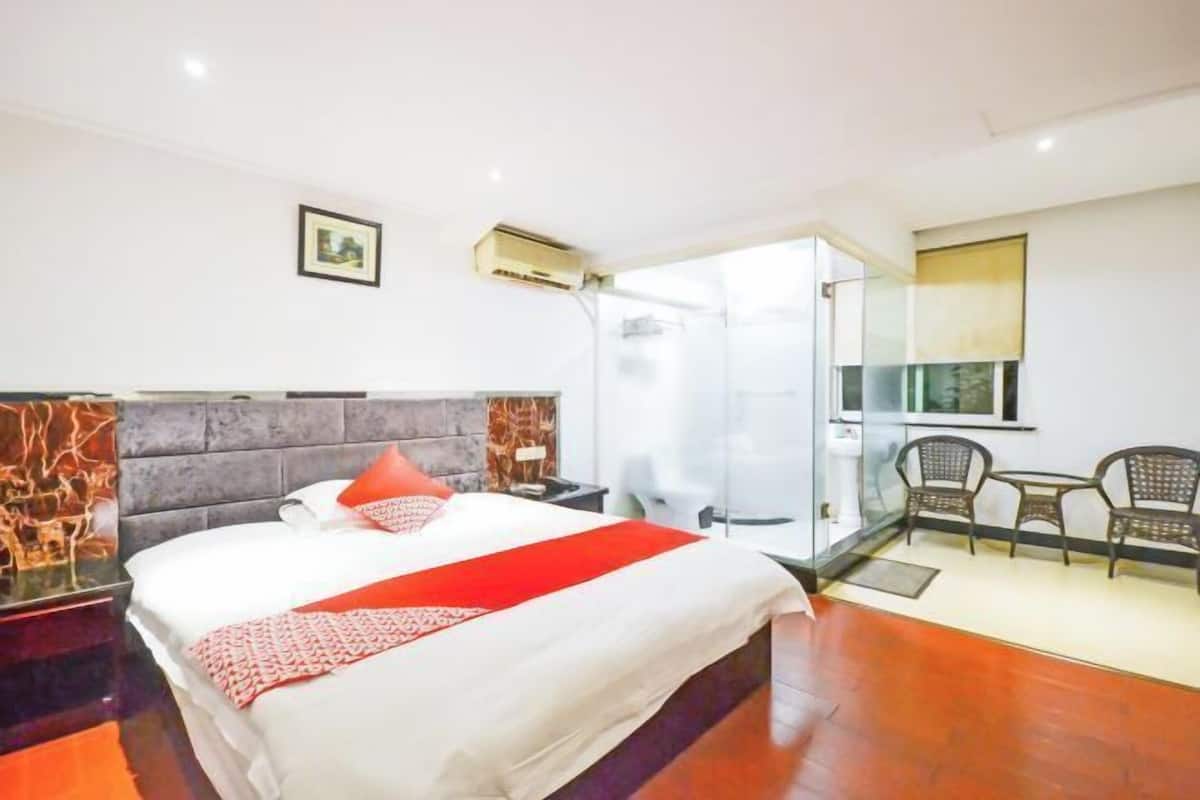 A hotel room with a bed, a nightstand, a chair, a small table, and a glass partition.