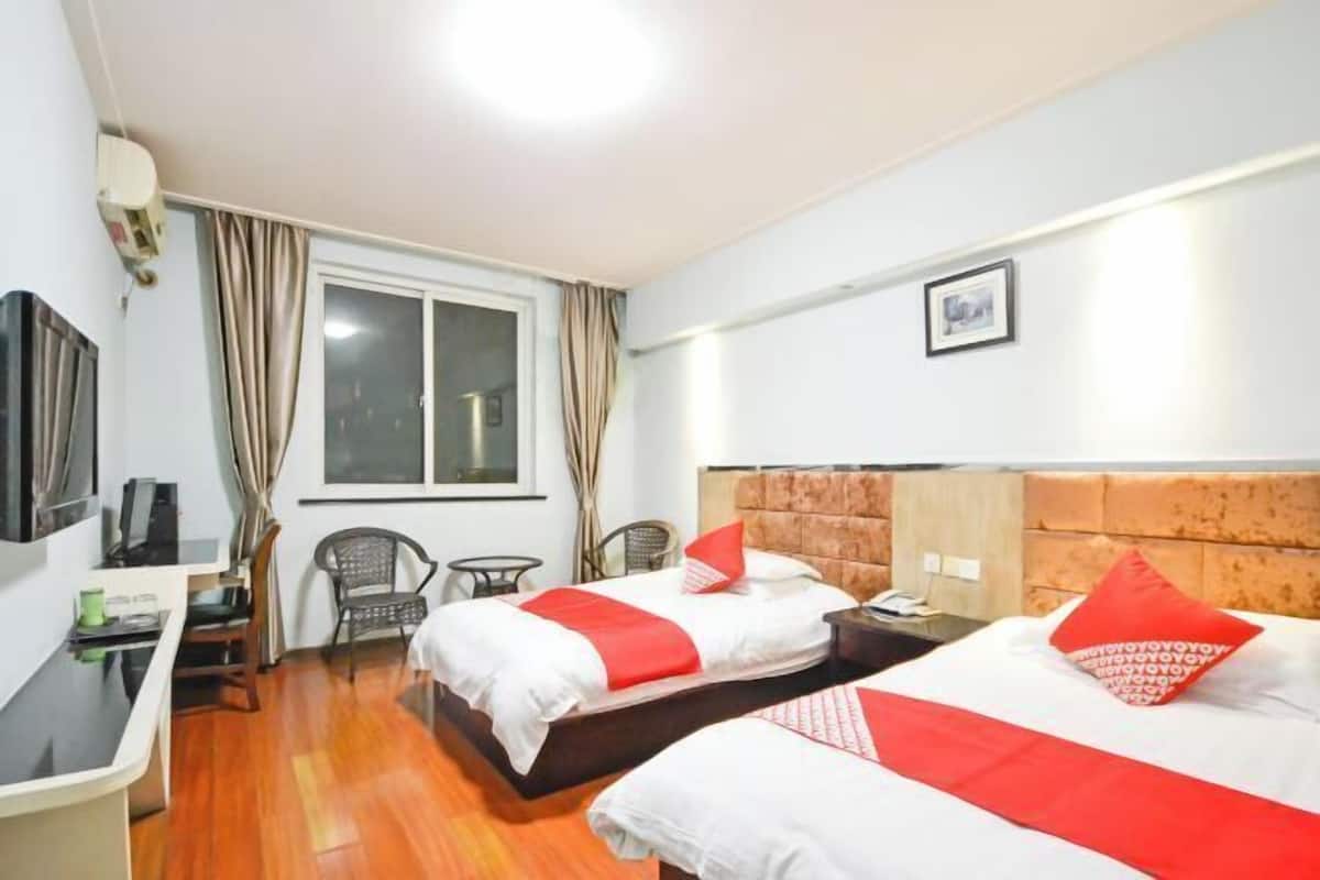 A hotel room with two beds, a desk, a chair, a window with curtains, and a framed picture on the wall.