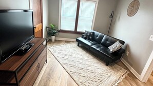 A living room with a flat-screen TV on a wooden stand, a black leather sofa, a beige rug, and a large window with blinds. - Modern Beach Retreat–Pet Friendly + Prime Location (Grand Haven)