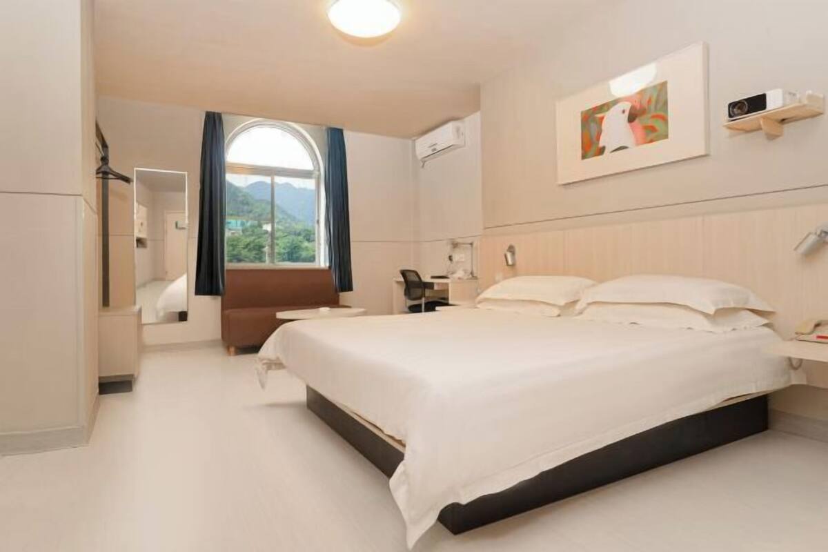 A hotel room with a bed, a desk, a chair, a window with curtains, and a painting on the wall.