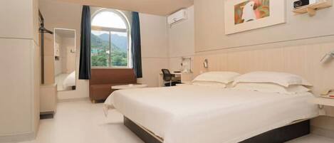 A hotel room with a large bed, a desk, a chair, a window with curtains, a mounted TV, and a framed picture on the wall.