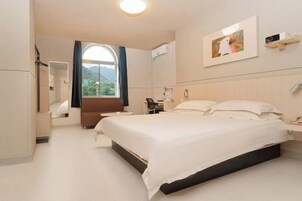 Habitación de hotel con una cama grande, un escritorio, una silla, una ventana con cortinas, un televisor montado y un cuadro en la pared.