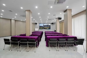 A conference room with multiple tables and chairs, a large screen at the front, and a view of a building through the windows. - Beijiang Inn (Qingyuan)
