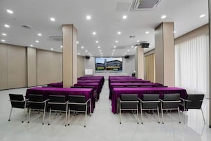 Un salón de conferencias con varias mesas y sillas, una pantalla grande al frente y vistas de un edificio a través de las ventanas.