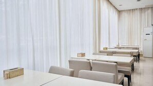 Een moderne eetruimte met witte tafels en stoelen, grote ramen met transparante gordijnen, en een strak, minimalistisch design.