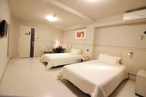 A hotel room with two beds, a bedside table with a lamp, a small desk, and a wall-mounted air conditioner.