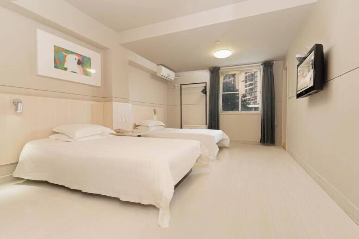 A hotel room with two beds, a window with curtains, a flat-screen TV mounted on the wall, and a ceiling light.