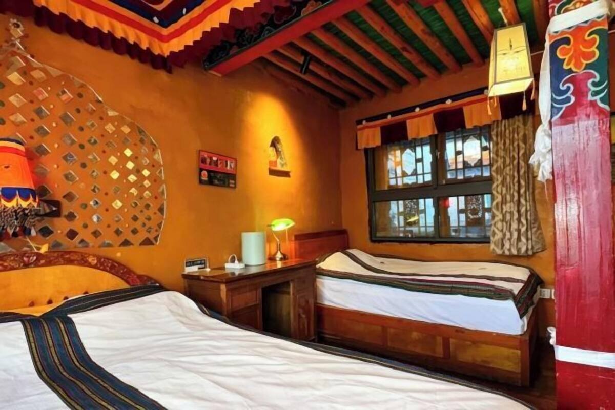 A room with two beds, a window with curtains, a wall-mounted lamp, and a decorative wall hanging.