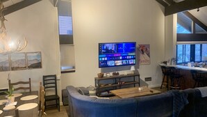 A modern living room with a curved blue sofa, a wooden coffee table, and a flat-screen TV mounted on the wall. There is a dining area with a round table and chairs, and a kitchen area with a bar counter and stools. - Spacious and Peaceful Location, Family and Pet Friendly (Mammoth Lakes)