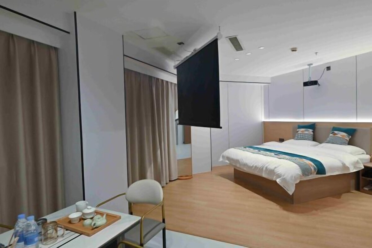 A modern hotel room with a large bed, a desk with a tray of items, and a TV on the wall.