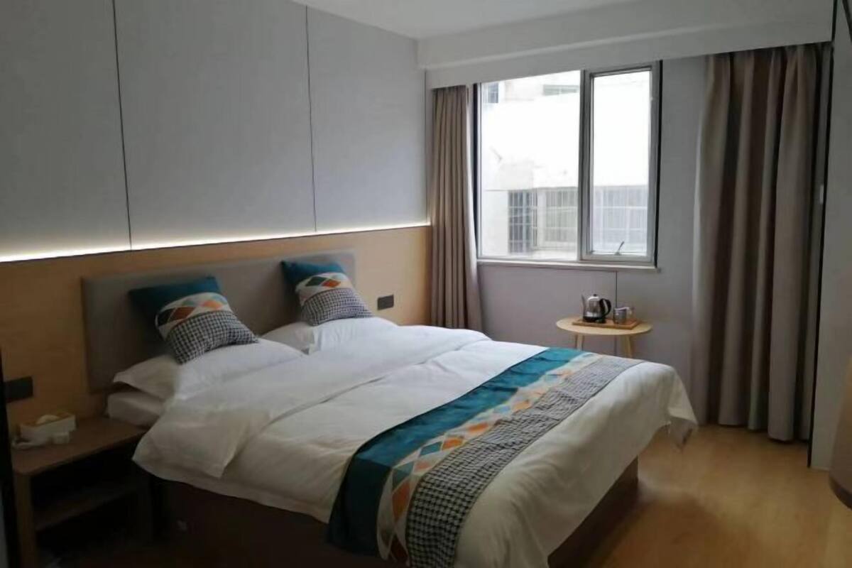 A hotel room with a bed, a nightstand, a small table with a kettle, and a window with curtains.