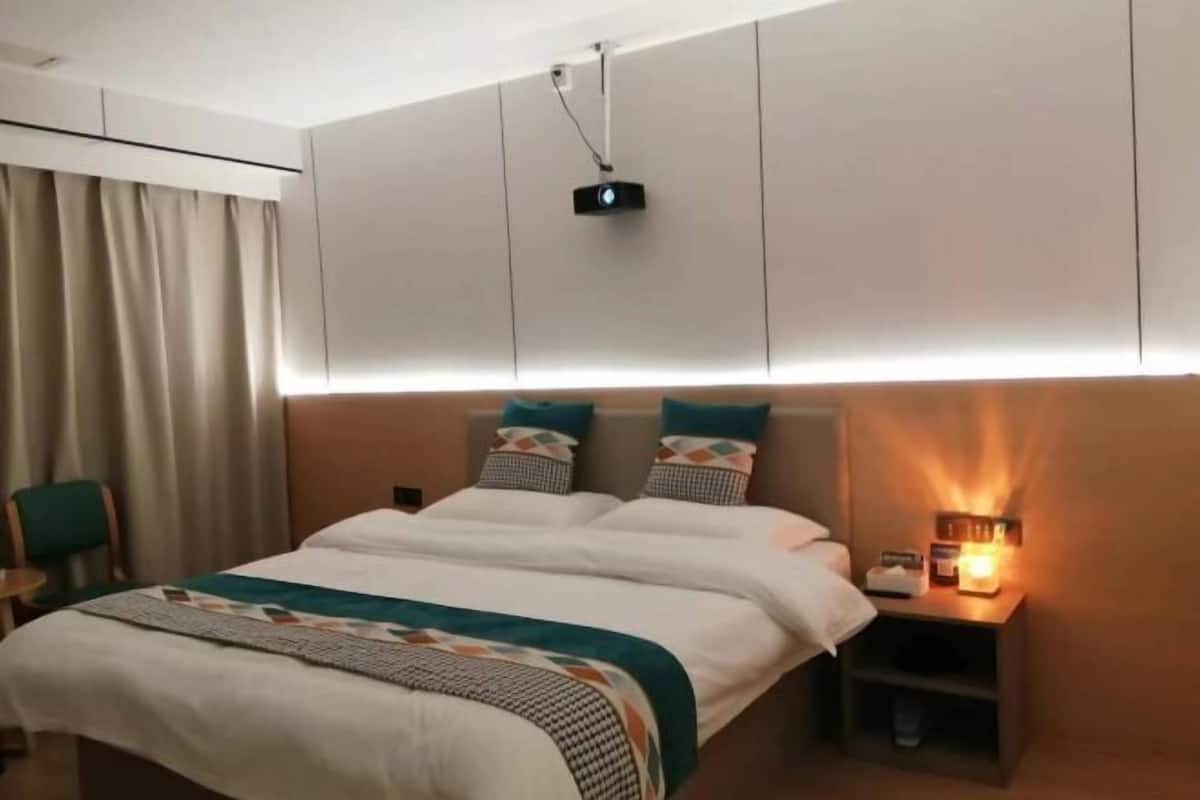 A hotel room with a bed, bedside table, lamp, and a projector mounted on the ceiling.